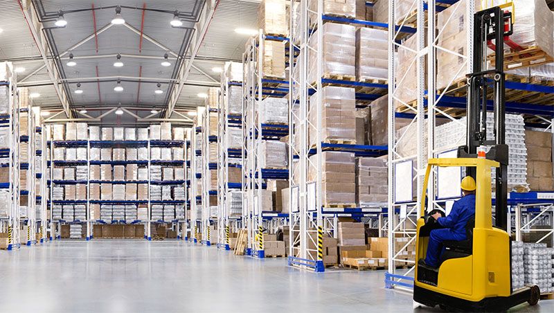 Warehouse interior with high shelves stacked with pallets and boxes, and a worker operating a yellow forklift to organize inventory.